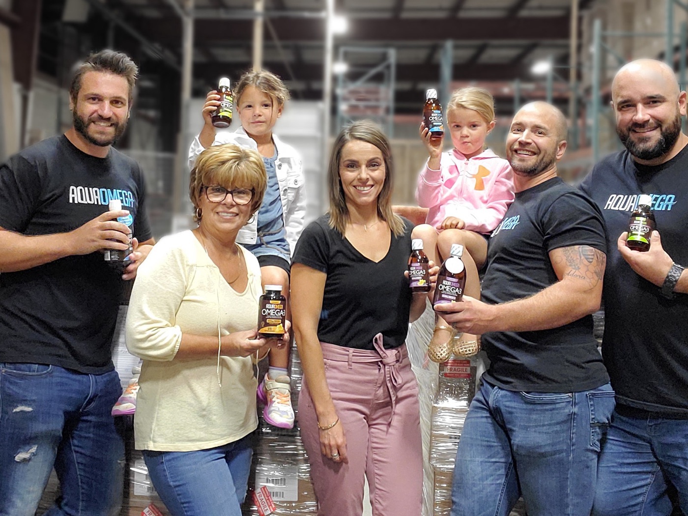 Some of the Vivo Brands team in the warehouse holding AquaOmega bottles, with two young helpers sitting on the pallets